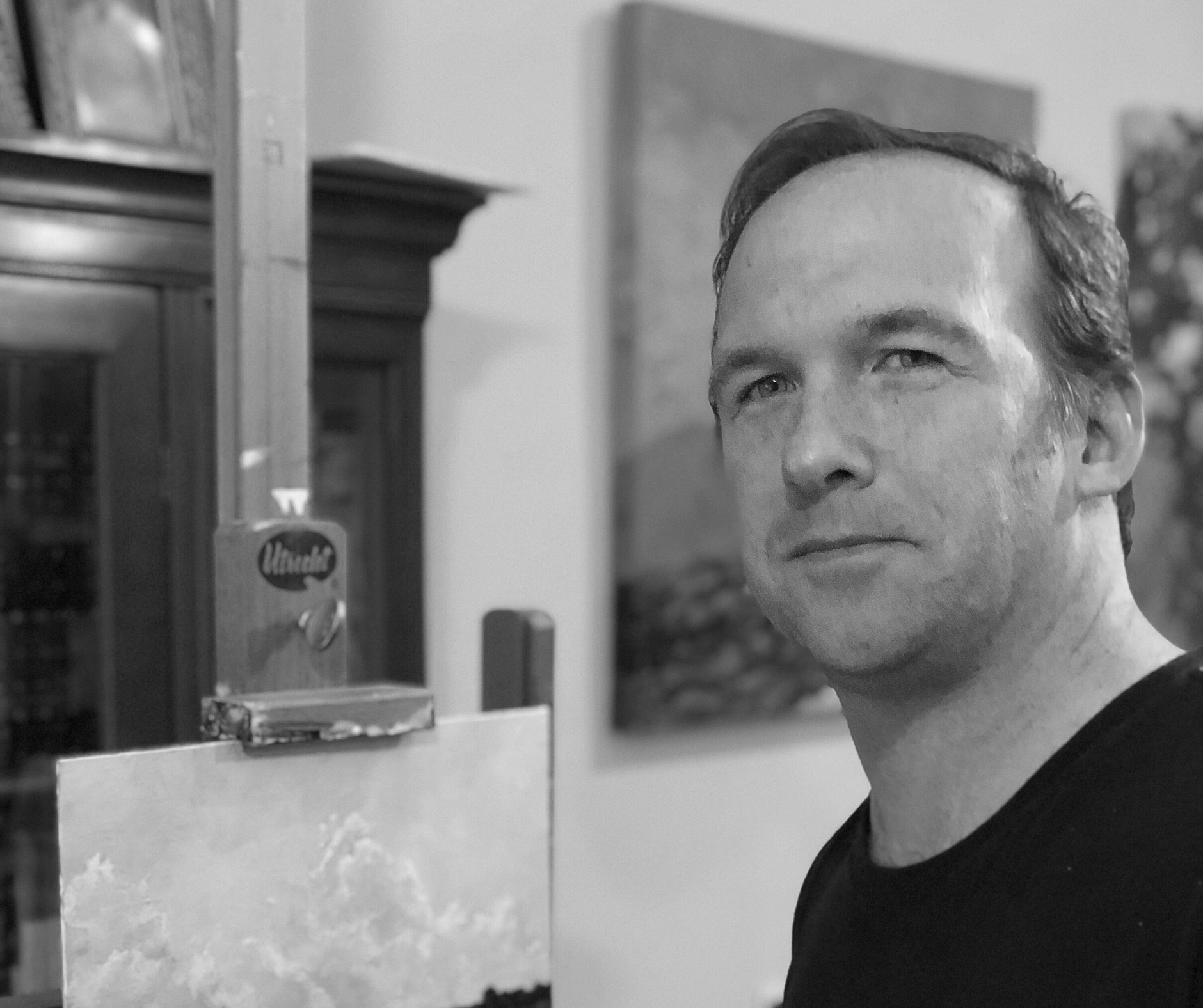 A Caucasian man, wearing a black shirt stands in front of his easel while looking back at the viewer. A landscape with clouds can be seen on his canvas.