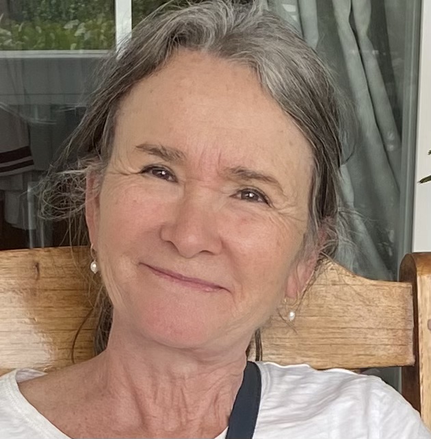 A woman with gray hair and pearl earrings sits on a wooden chair, smiling slightly, with a white curtain and greenery in the background.