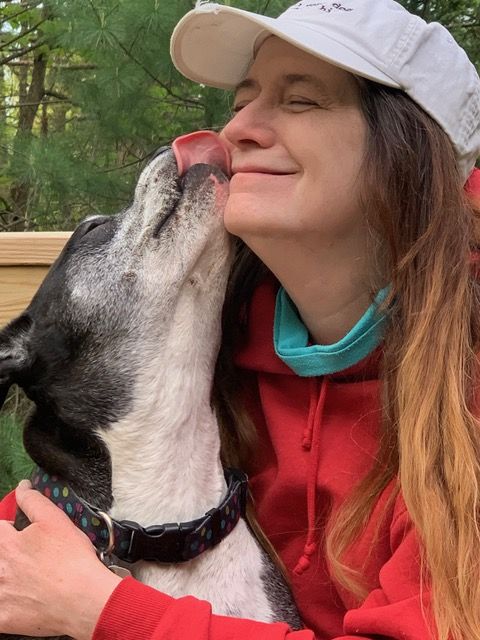 A woman in a red hoodie and white cap smiles as a black and white dog licks her face outdoors.
